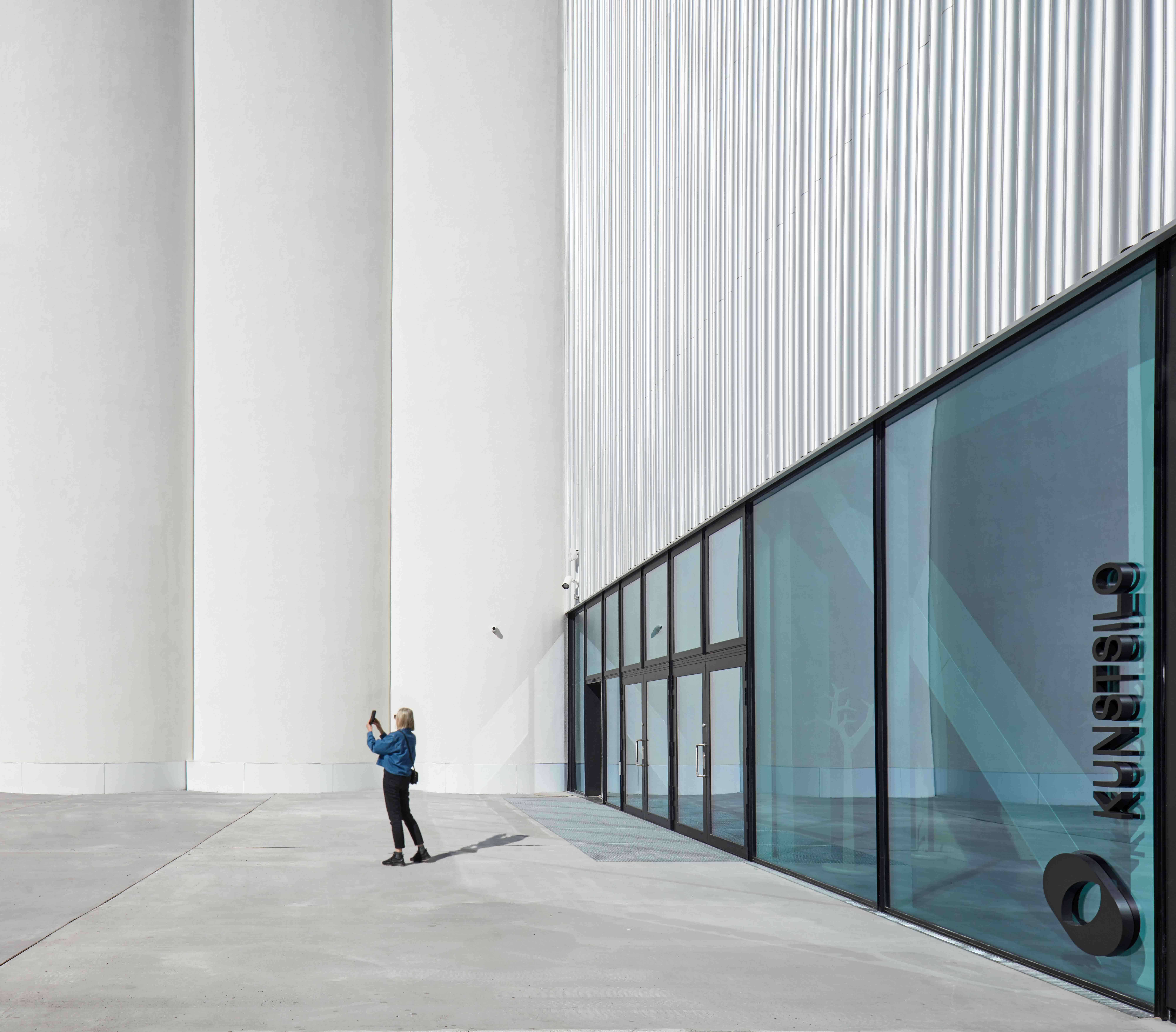 En kvinne står utenfor en monumental betongbygning og tar et bilde med mobiltelefonen.