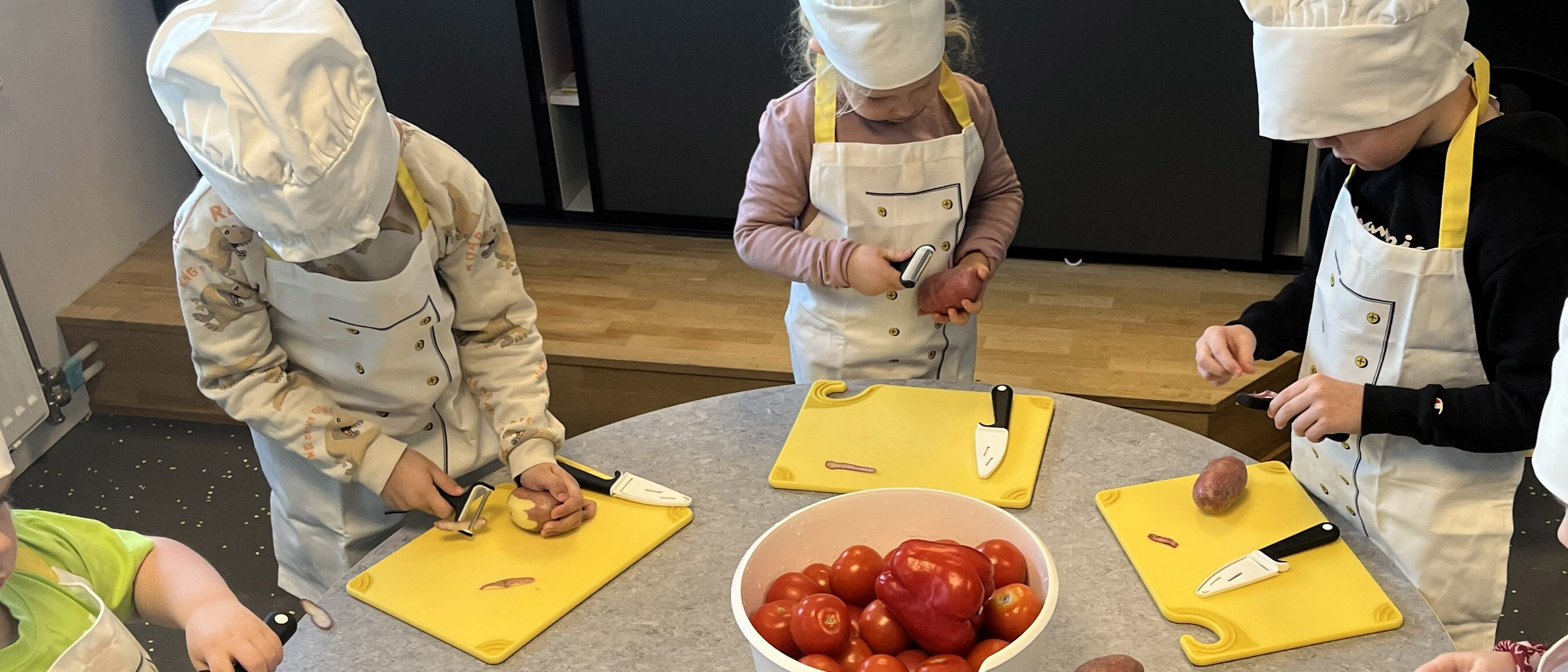 Barn med kokkeluer lager mat ved et rundt bord.