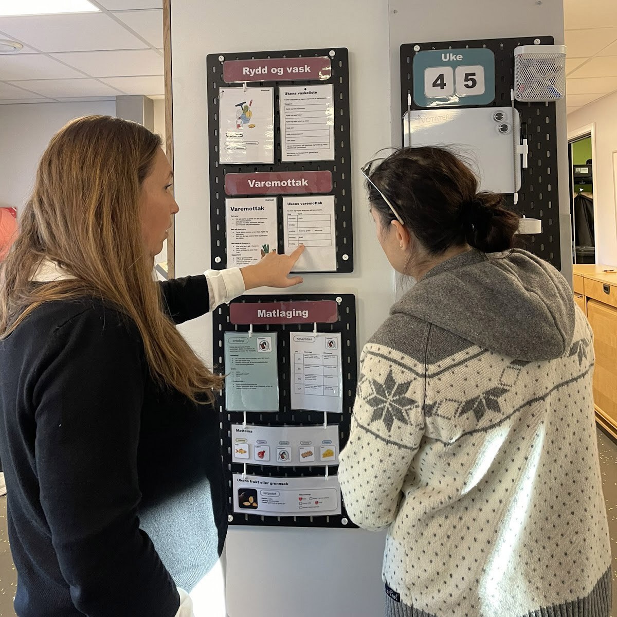 To kvinner står og diskuterer en arbeidsplan som henger på en vegg.