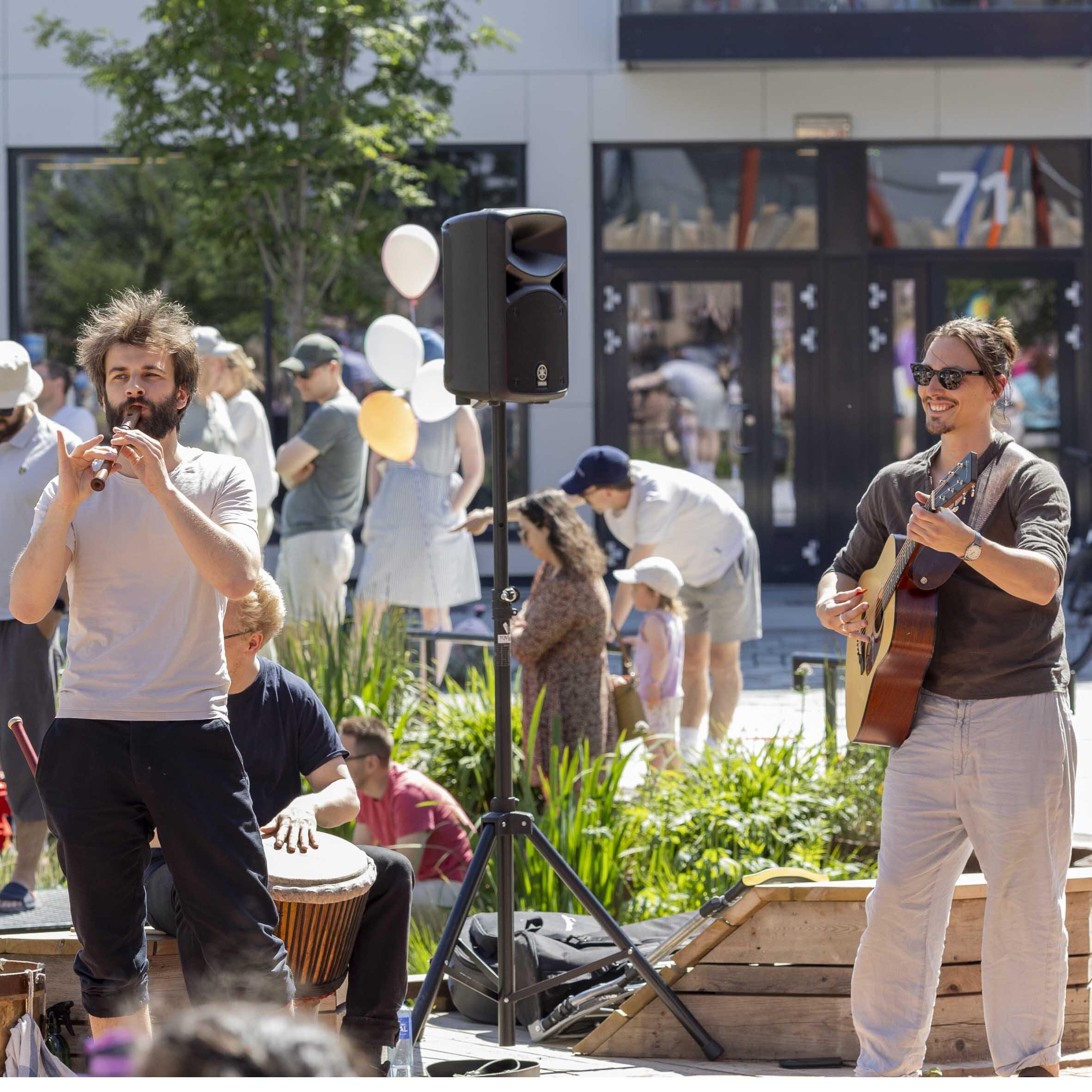 Musikere opptrer på et torg, med høyttalere og publikum i bakgrunnen. Rundt dem står voksne og barn samlet i solskinnet, med bygninger og grøntområder bak.