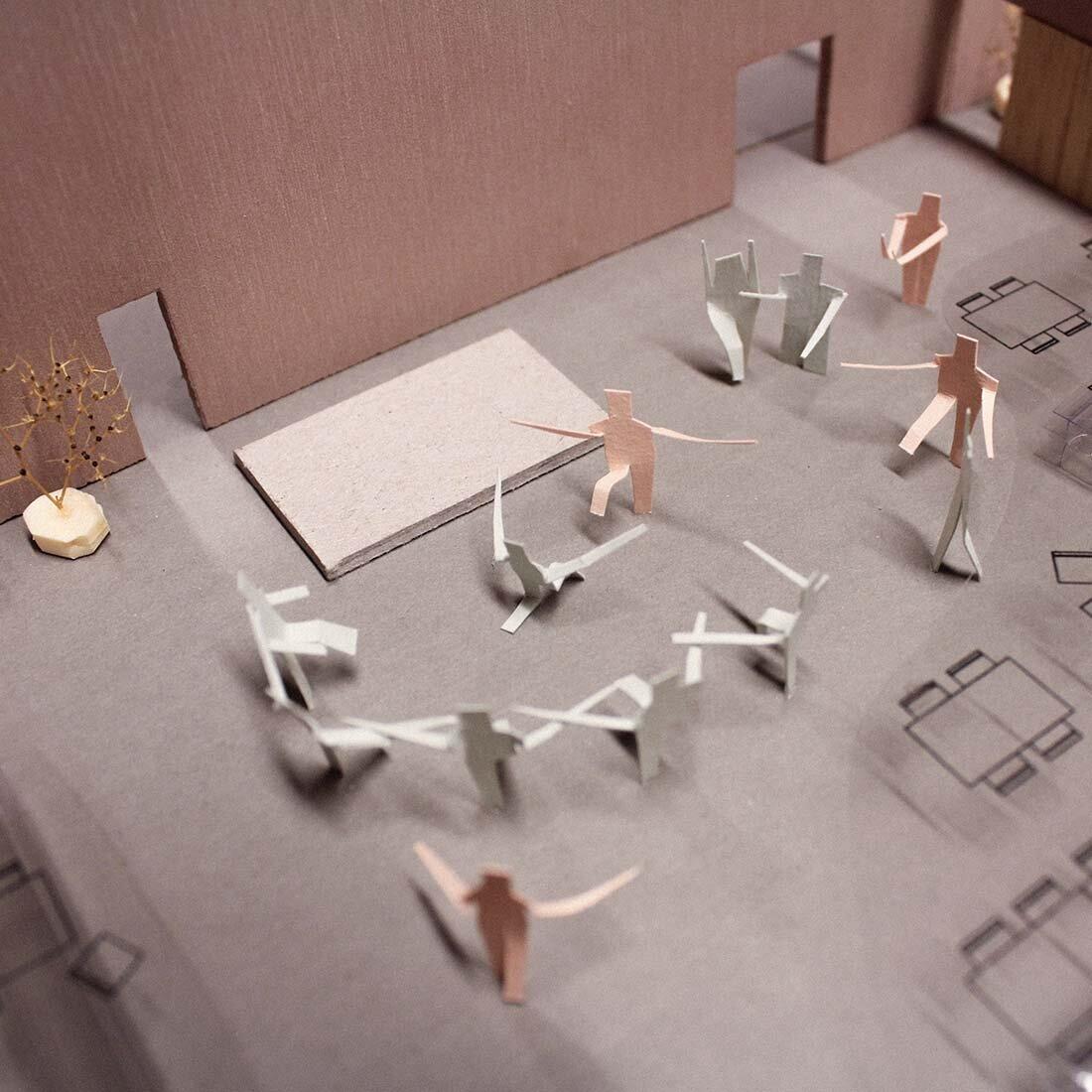 Vi ser ned i en pappmodell av et forsamlingsrom der papirmennesker ser ut til å danse sammen.