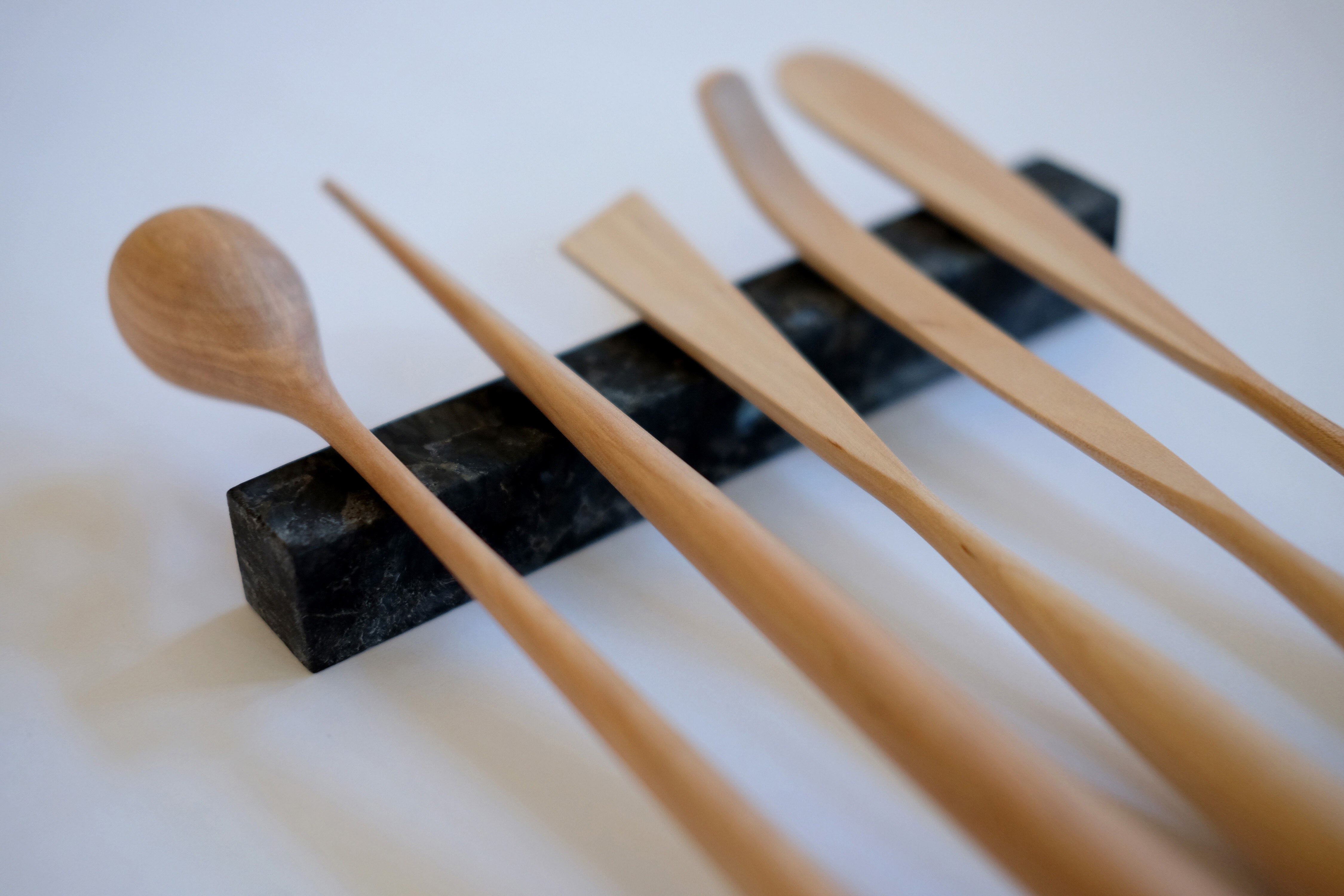 En serie på fem bestikkdeler i lyst treverk er plassert med spiseenden på en lang sort bestikkholder.