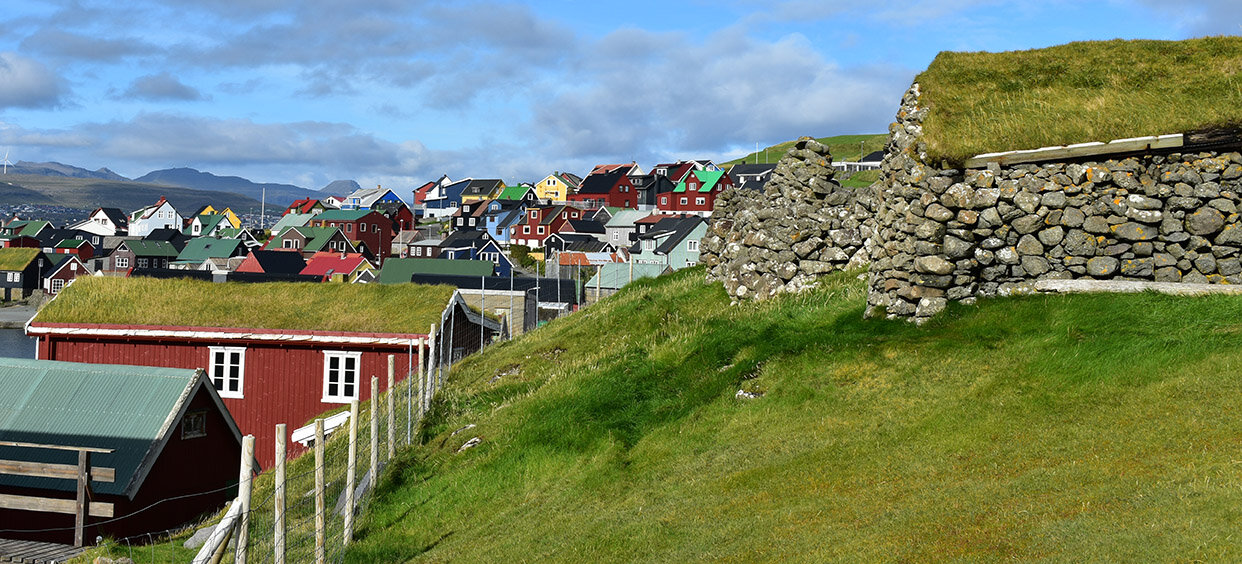 Et oversiktsbilde over et Færøysk landskap med slette og gamle steinhus i forgrunnen og fargerik trehusbebyggelse i bakgrunnen.