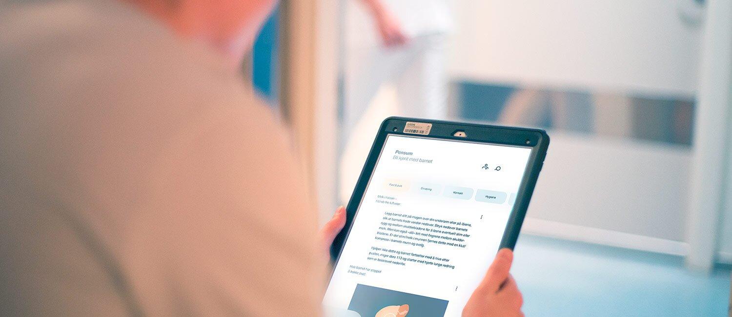 Bildet viser en person som holder et nettbrett. Det er kun nettbrettet som er i fokus, men en skimter kliniske omgivelser.