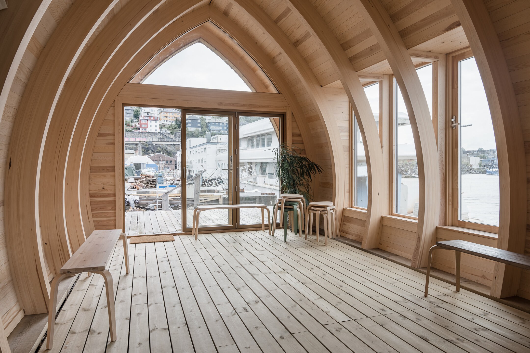Et rom med høyreiste, bueformede limtrebjelker. Endeveggen er vinduer og en skyvedør i glass.