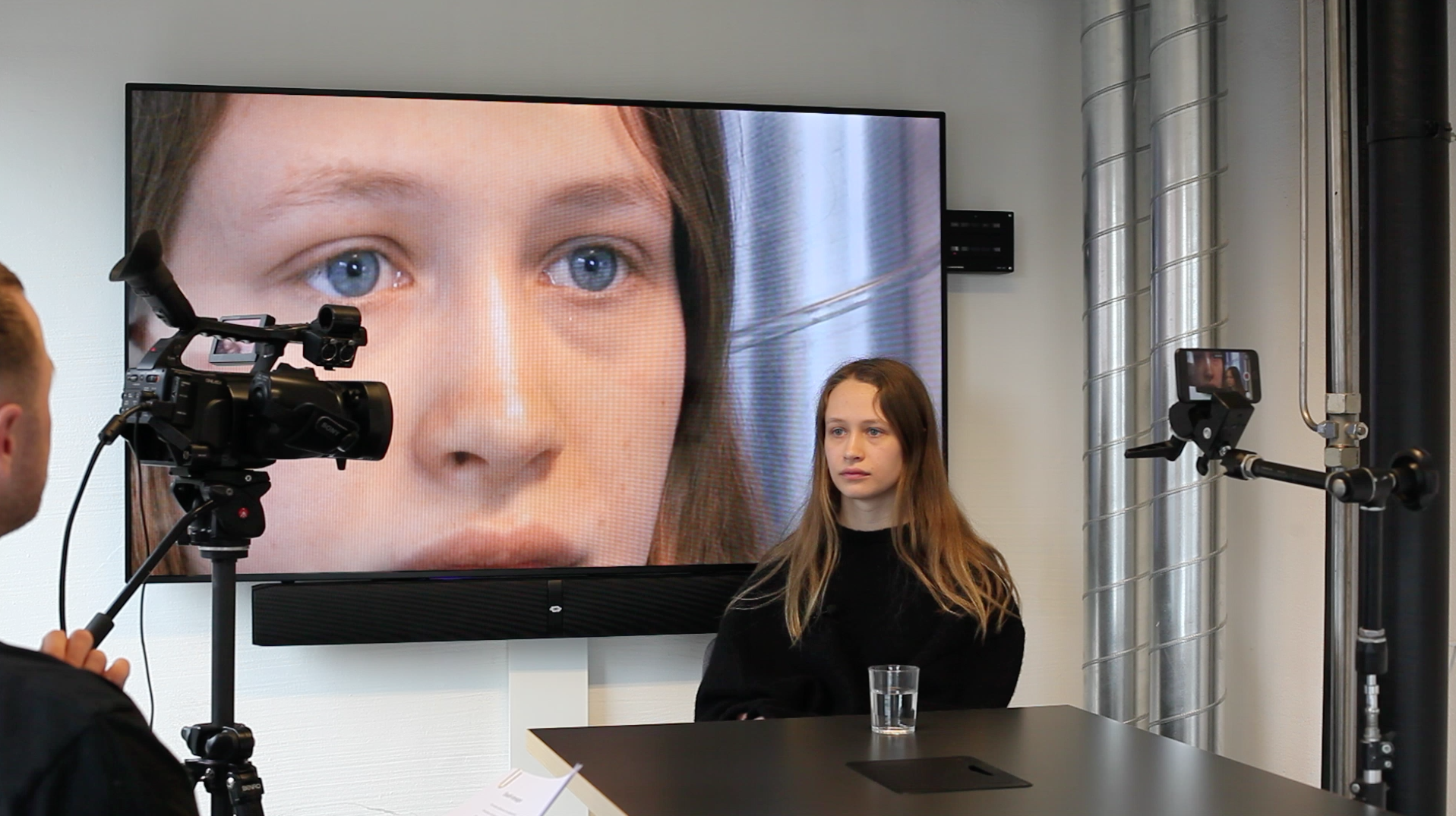 Bilde av en ung kvinner som blir filmet. Hun sitter ved et bord, og bak henne ses et nærbilde av ansiktet hennes på en skjerm.