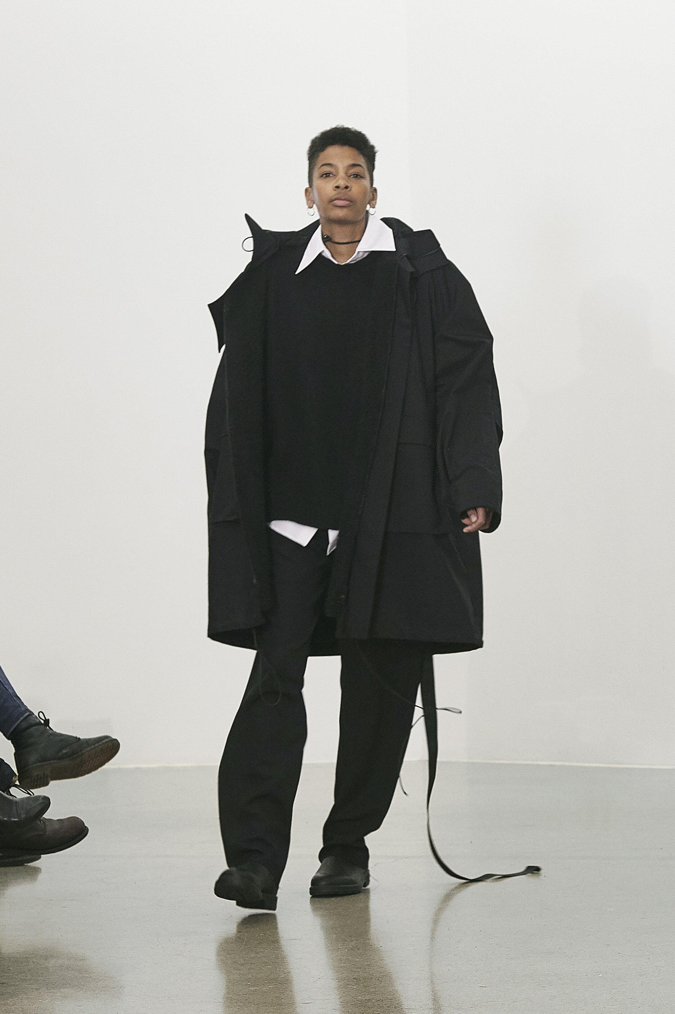 Bilde av en person i et romslig sort antrekk med hvit skjort i bunn, som går på en catwalk.