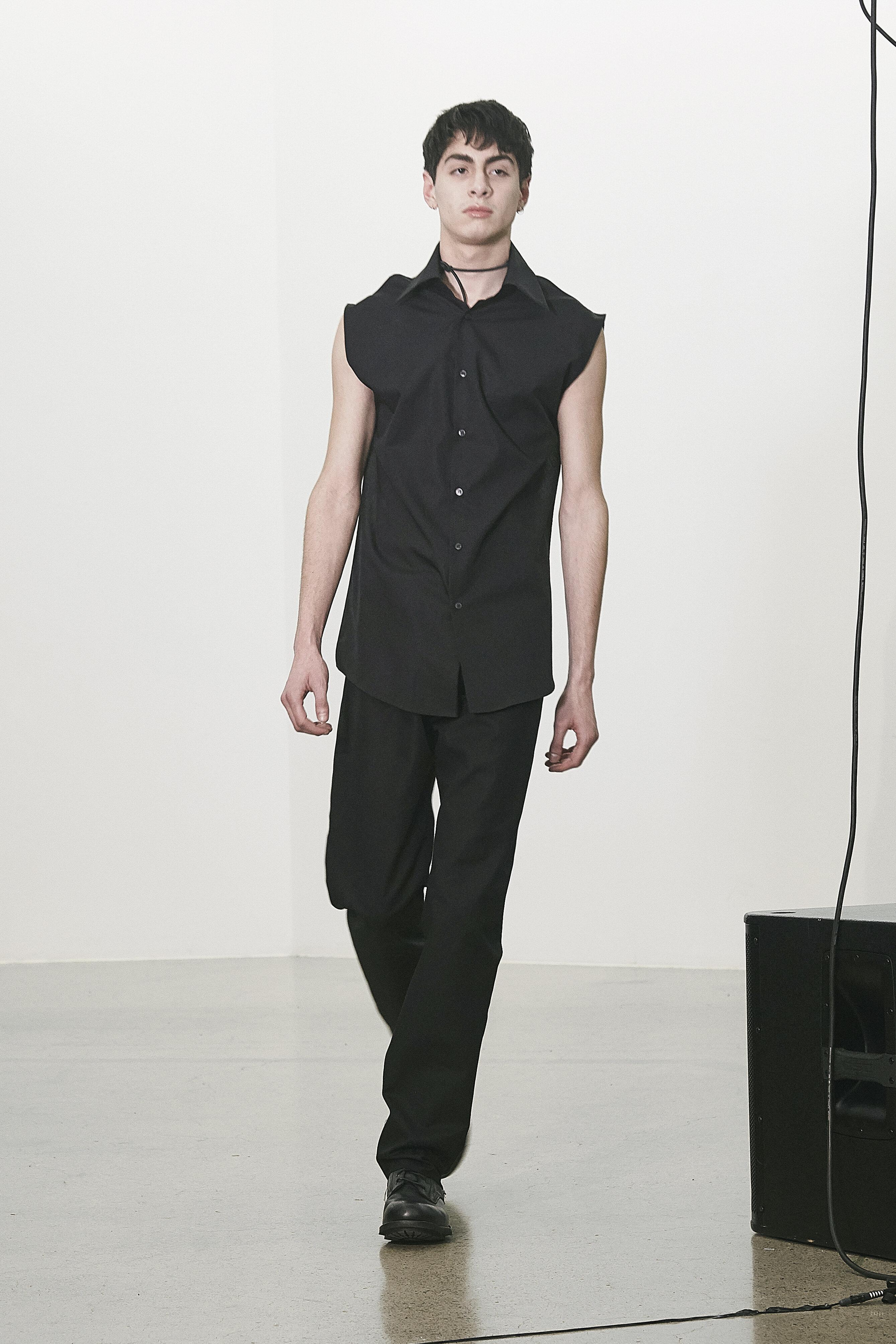 En slank høy mann ikledd et sort ermeløst antrekk går nedover en catwalk.