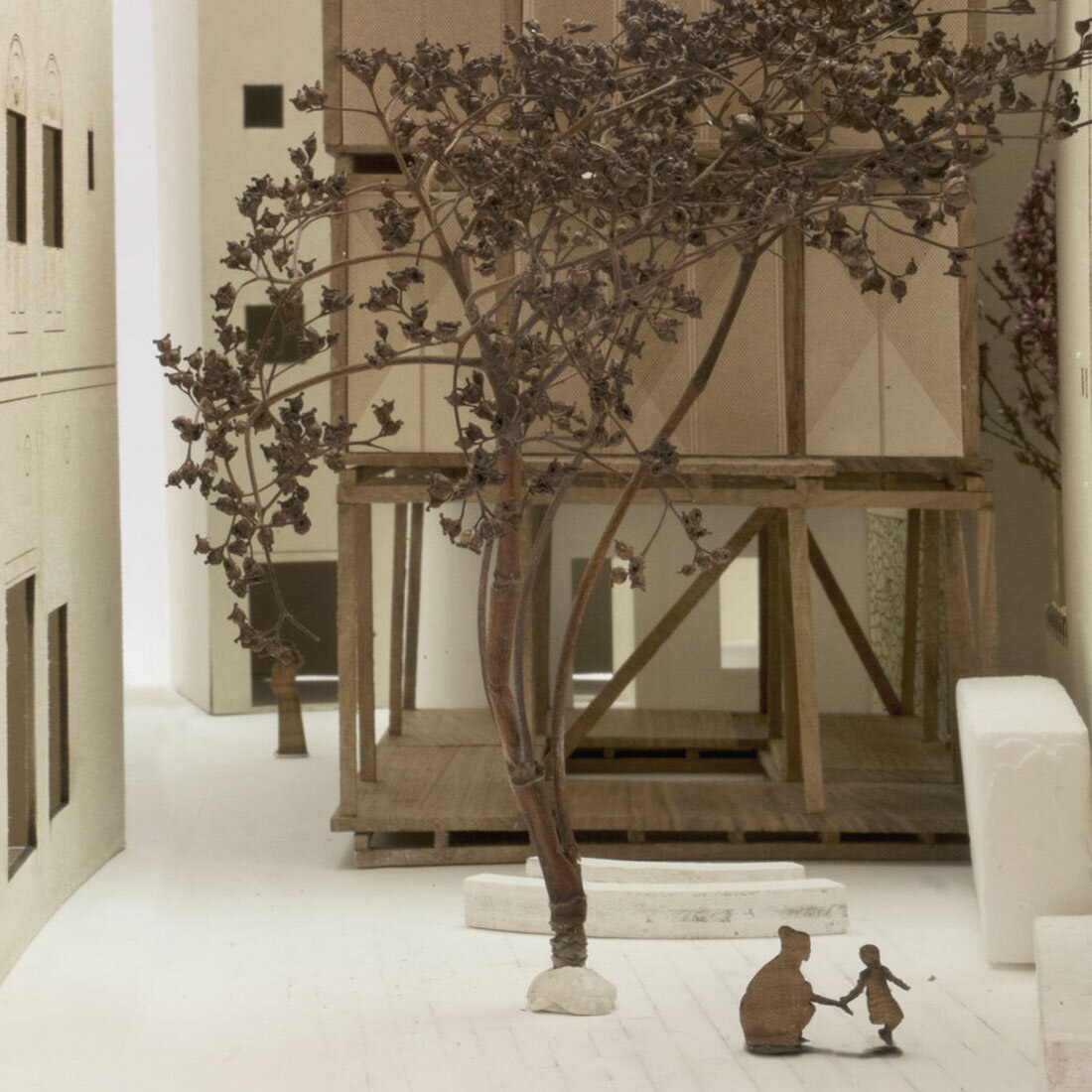 Et bilde inni en fysisk arkitekturmodell som viser en kvinne og et barn på en gårdsplass omringet av høy bebyggelse og et stort tuntre.