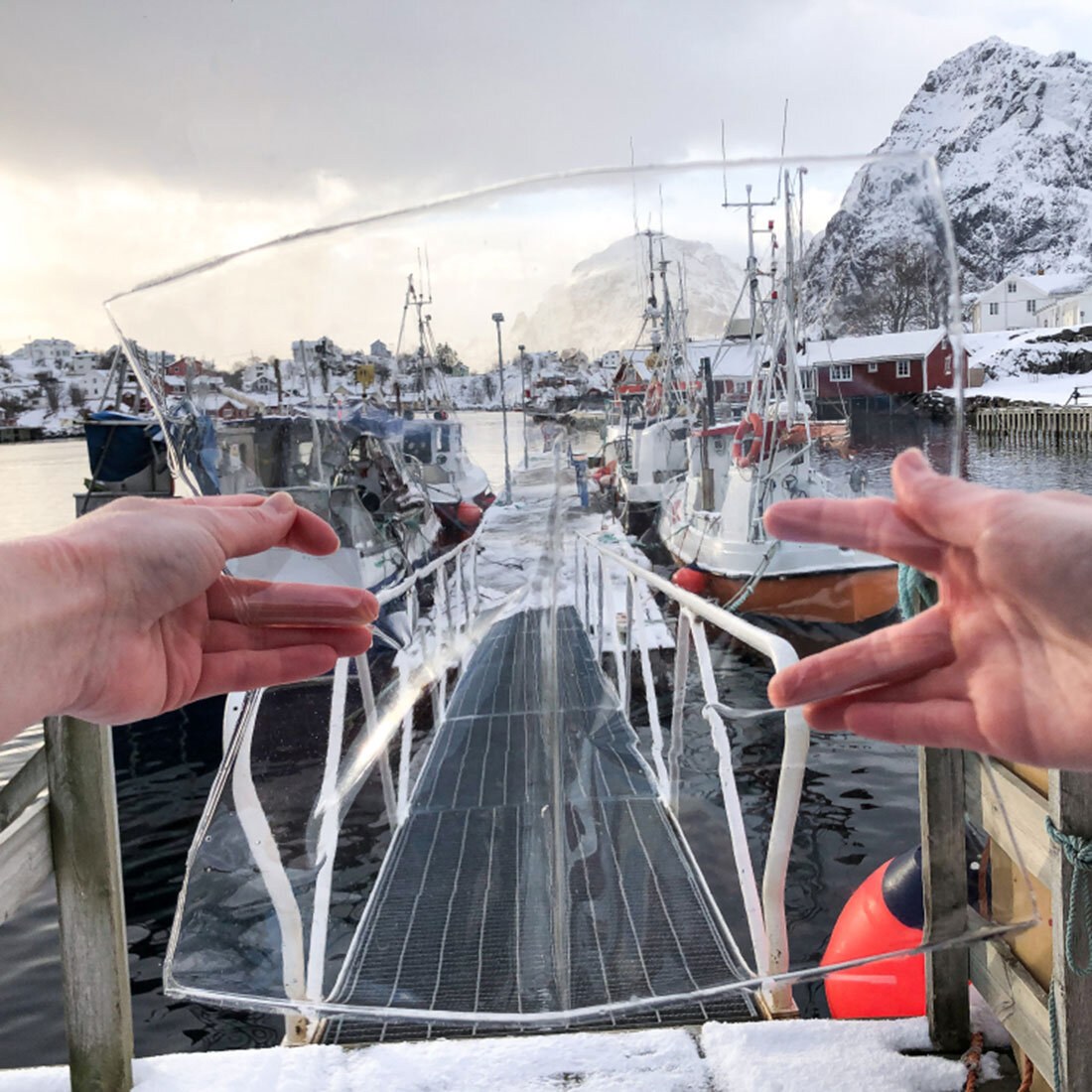 To hender holder opp et gjennomsiktig panel som vi se omgivelsene gjennom: en kai med fortøyde fiskebåter og snødekte fjell.
