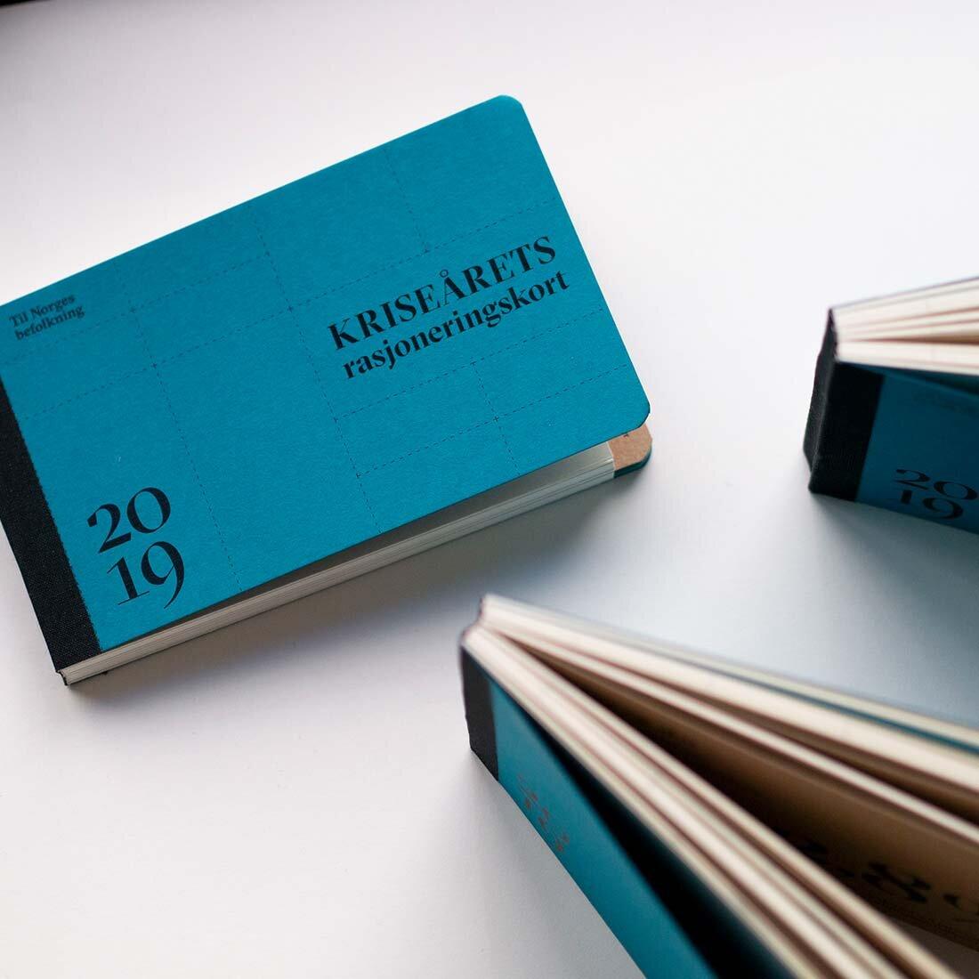 Blå bok på hvitt underlag. På boken står det: "Kriseårets rasjoneringskort" og "2019"