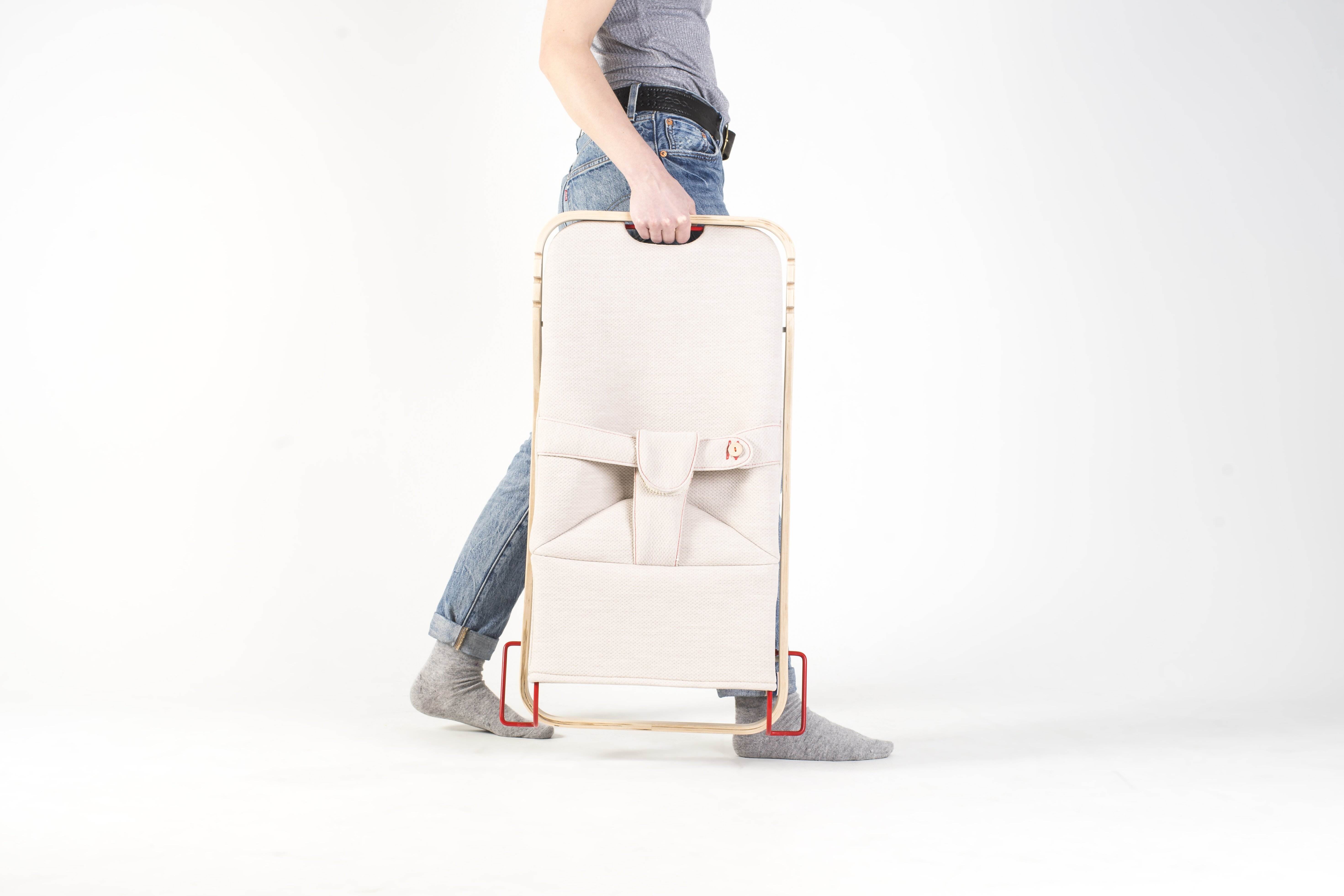 Bildet viser en person som holder en sammenlagt vippestol. Stolen har stativ i naturlig treverk, lyst tekstil og ramme og detaljer i rødt metall.