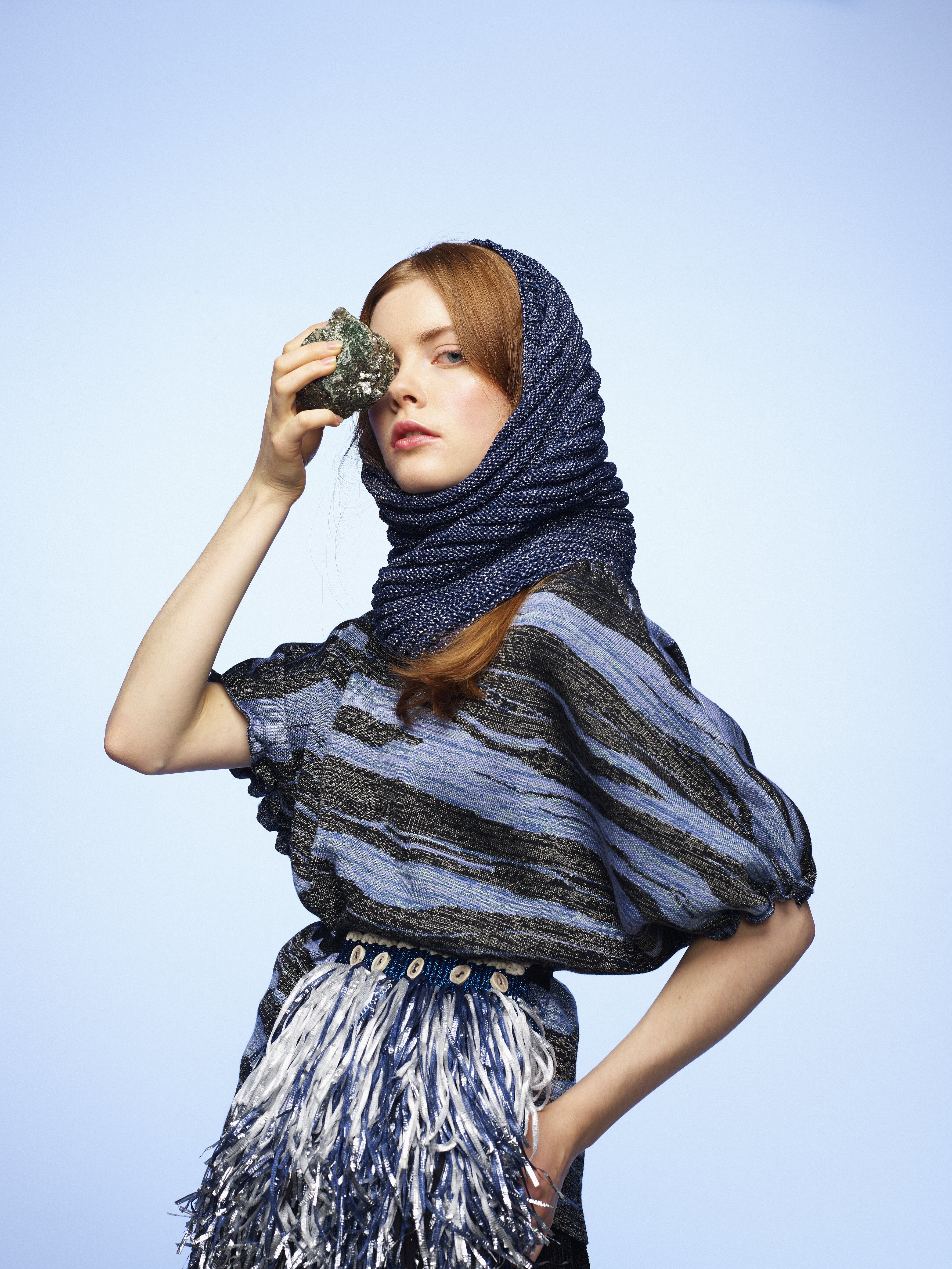 En kvinne med i en vevd mønstret kjole og sjal inspirert mot en lys bakgrunn. I livet har hun et belte med lange frynser.