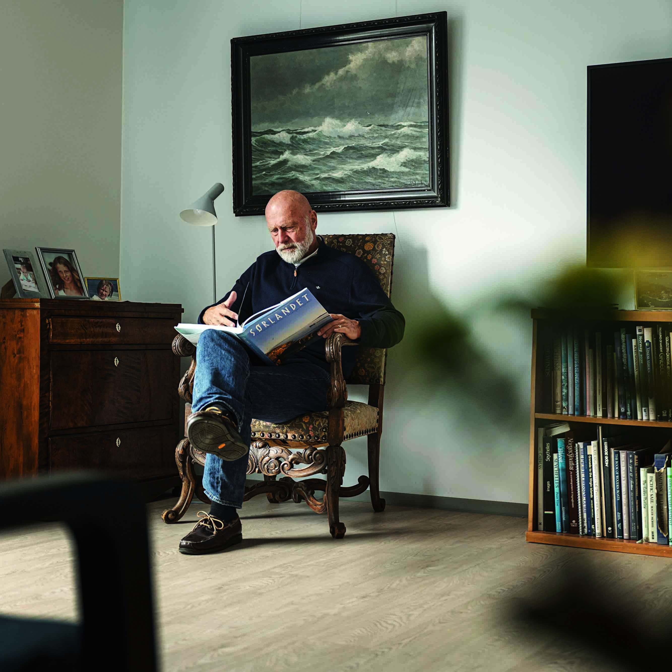 En eldre mann sitter i en stue og leser i en bok.