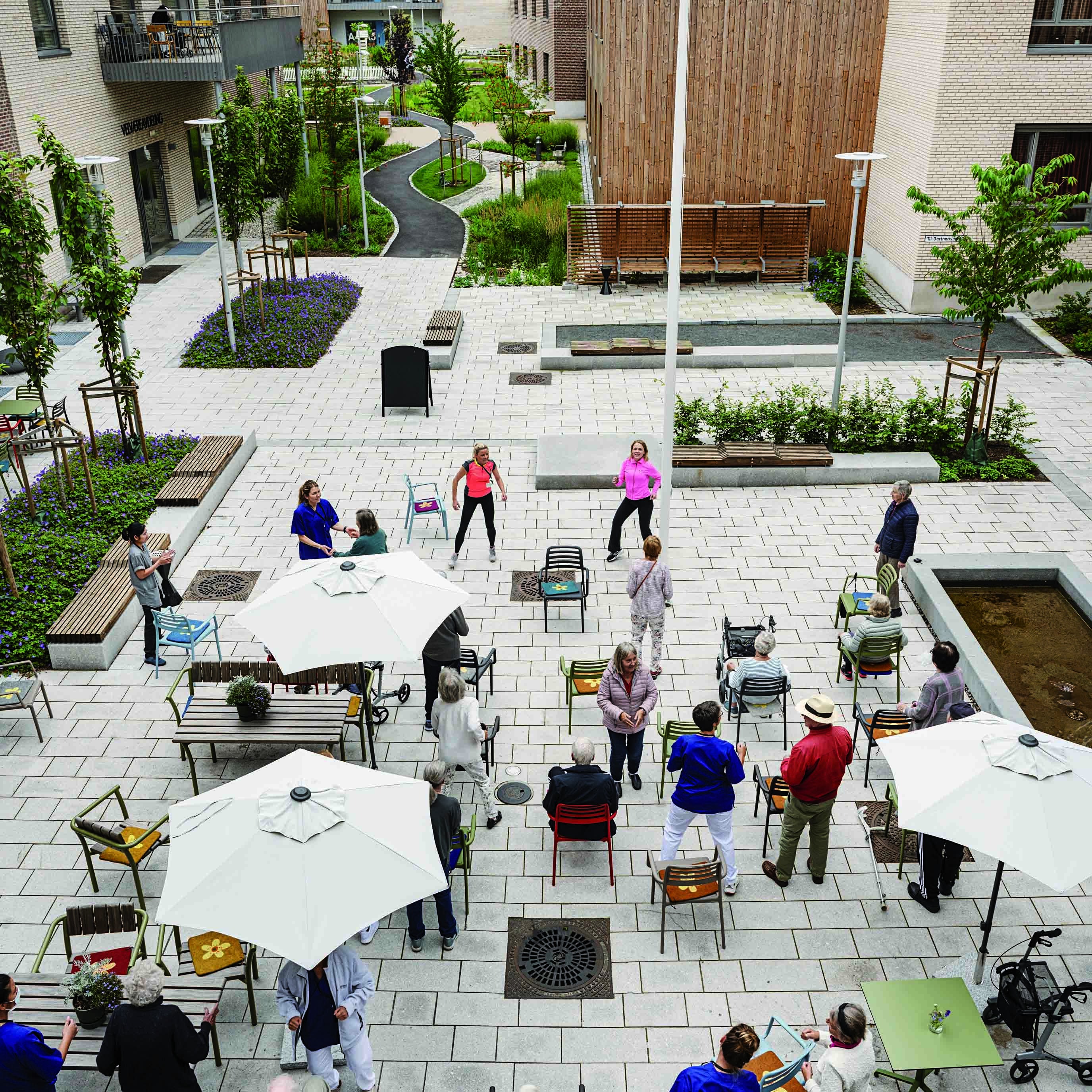 To treningsinstruktører instruerer en gruppe eldre som står eller sitter på stolene sine ute på en hellelagt åpen plass.