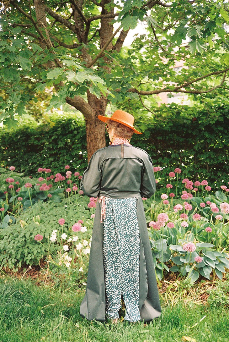 Eldre dame står ute i en hage, kledd i mørk og lang kjole og oransje hatt.