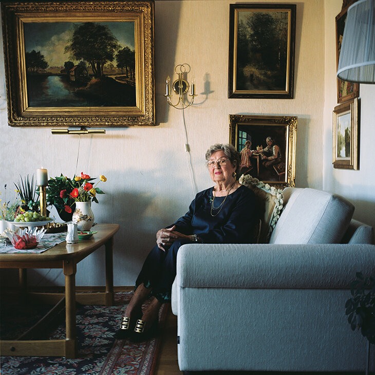 Eldre dame sitter smilende vendt mot kamera i en sofa.