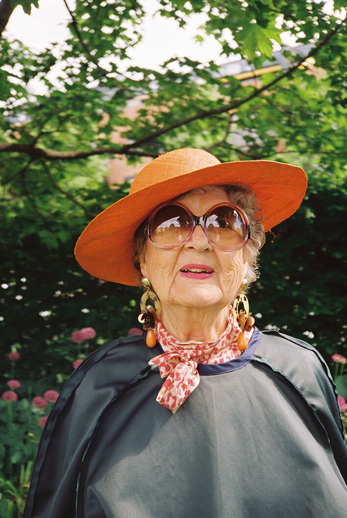 Nærbilde av eldre dame med solbriller, oransje hatt og mørk kjole.