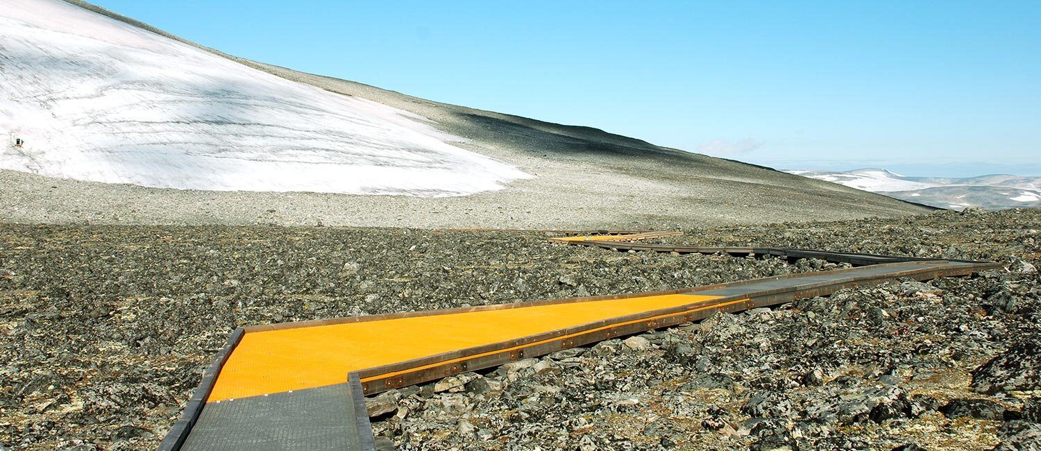 En gangsti i metall gjennom et røft steinlandskap med myke, isdekte fjell i bakgrunnen.