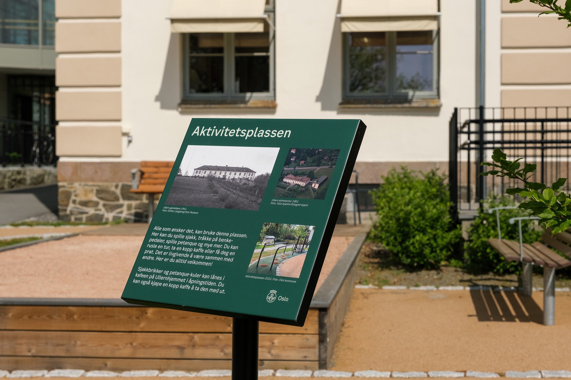 Grønt informasjonsskilt ved aktivitetsplassen i parken, med benk og uteområde i bakgrunnen.