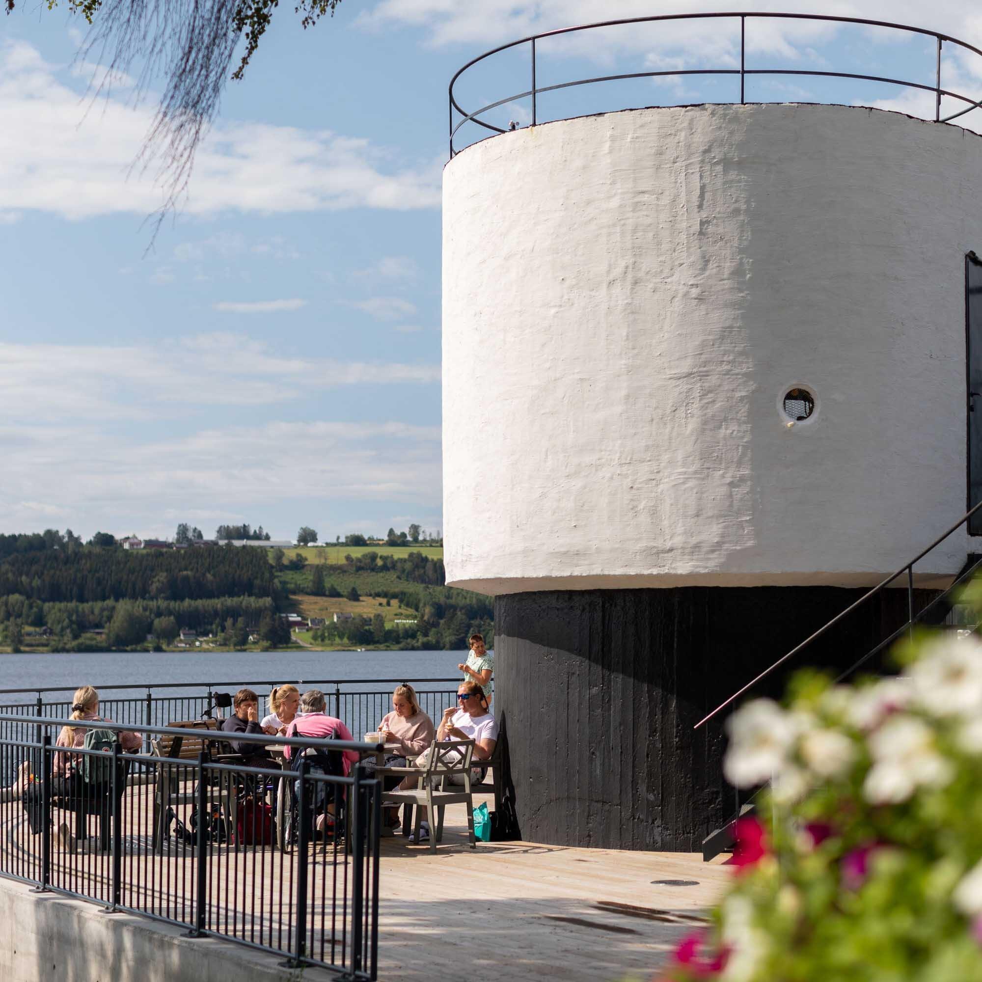 En sylinderformet betongkonstruksjon, et pumpehus. Nederst en terrasse der det sitter folk og spiser is