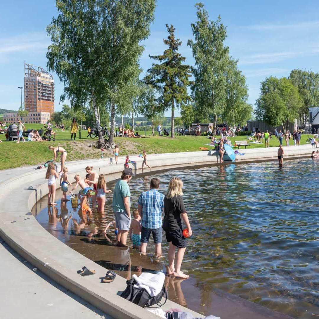 Strandpromenade i betong. Voksne som vasser i vannet, barn som leker og bader i vannkanten.