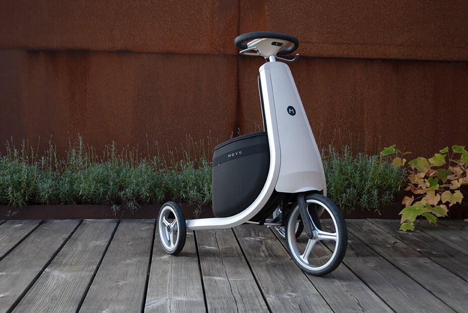 Hvit trehjuling Movu med ratt mot rustrød vegg, sett fra siden.