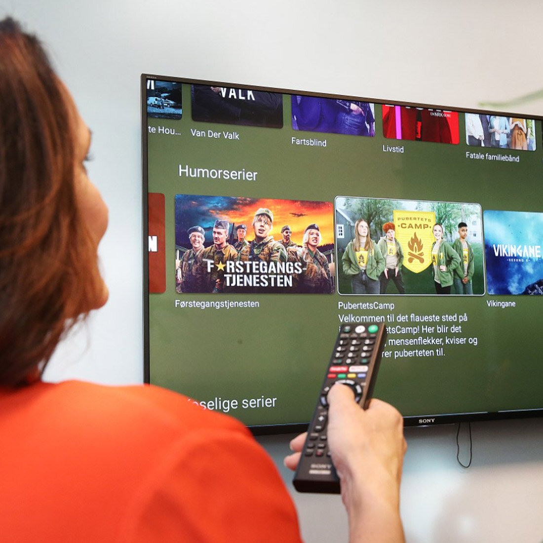 En kvinne peker på en smart-tv med en fjernkontroll. På skjermen ses en programvelger i NRKs app for smart-tv.