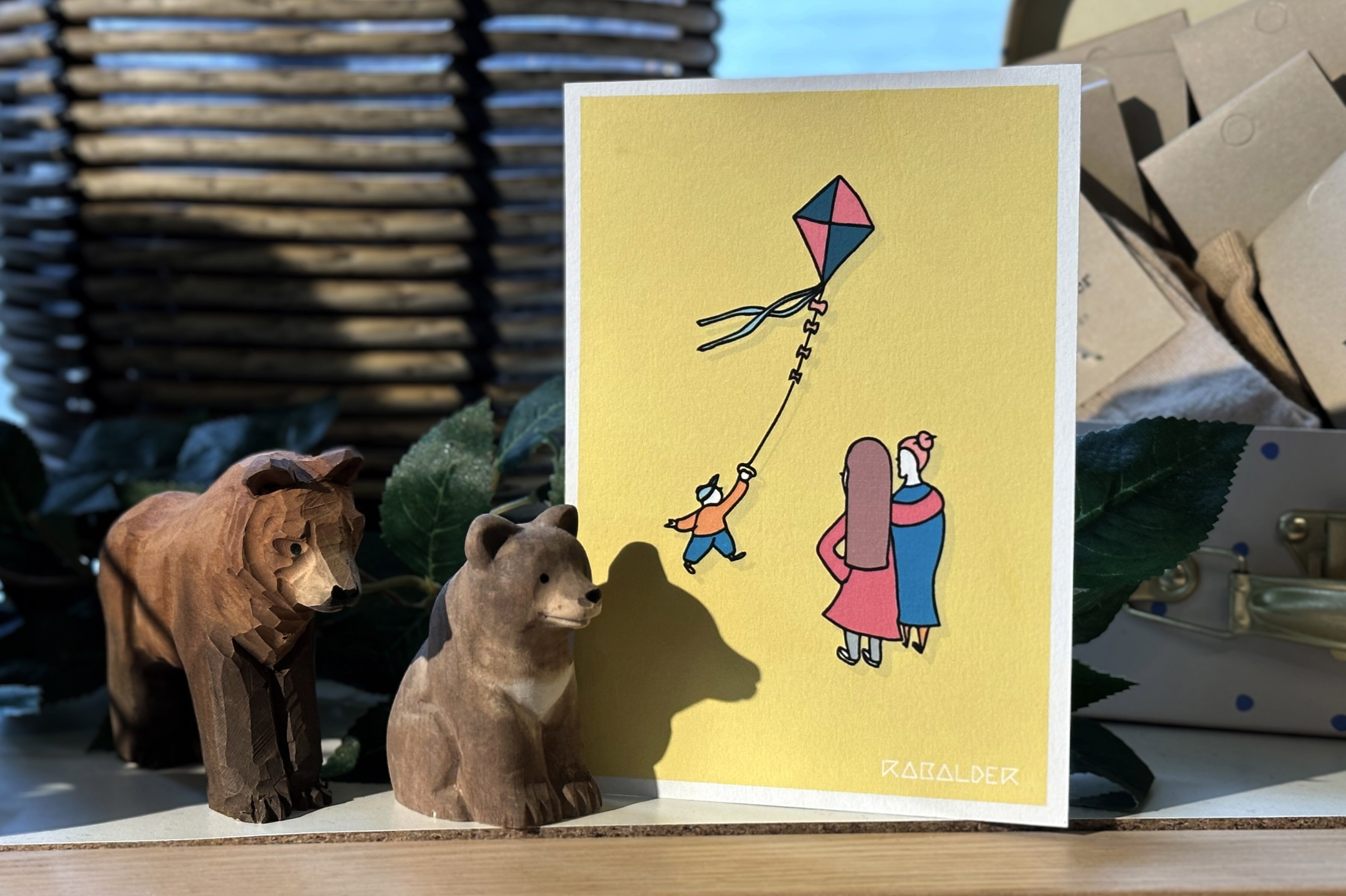 Illustrert gratulasjonskort der en gutt flyr en drage mens to kvinner står ved siden av og ser opp.