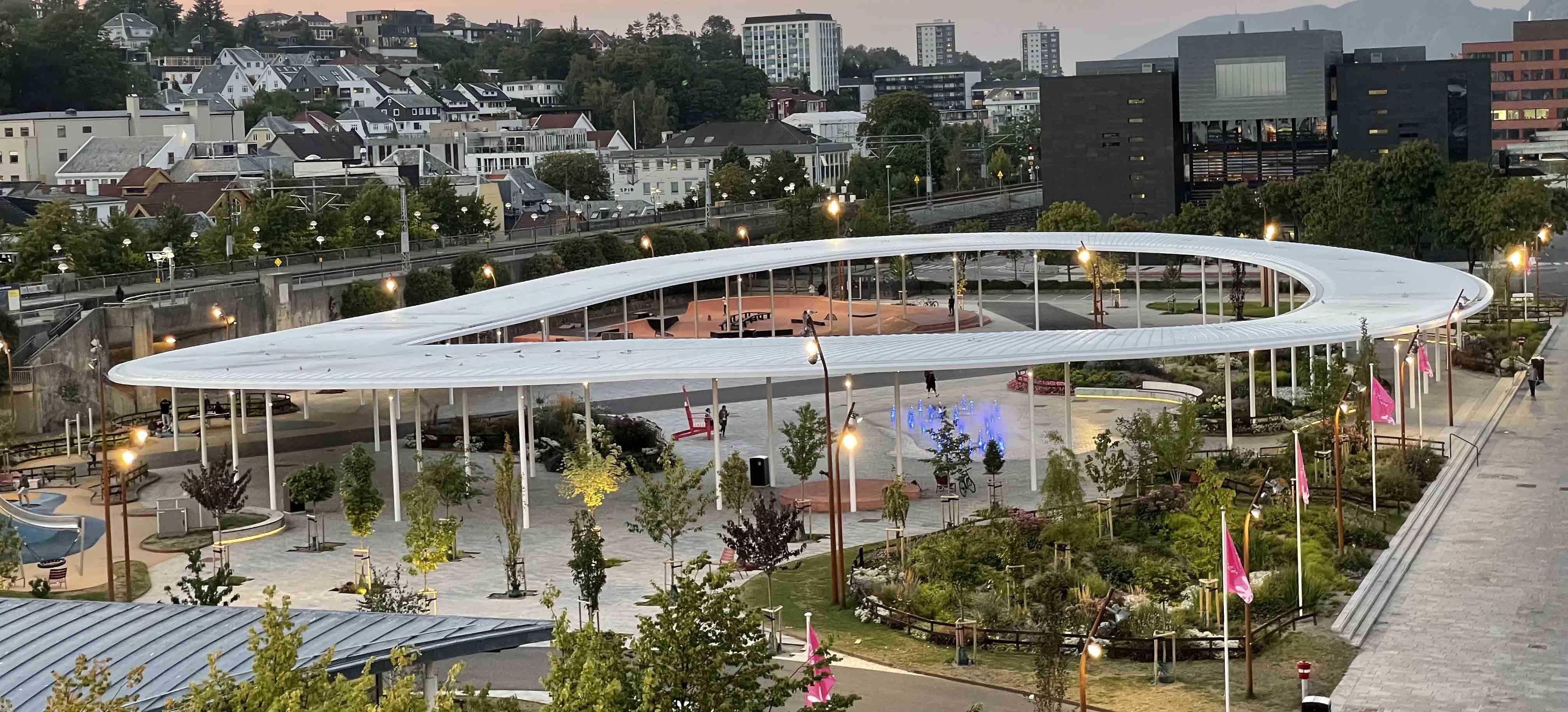 Et stort, bølgende, rundt tak, åpent i midten, over en steinlagt plass med bed og beplantning.