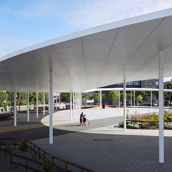 En person triller en annen i rullestol over et betongdekke under et sirkelformet tak holdt oppe av søyler.