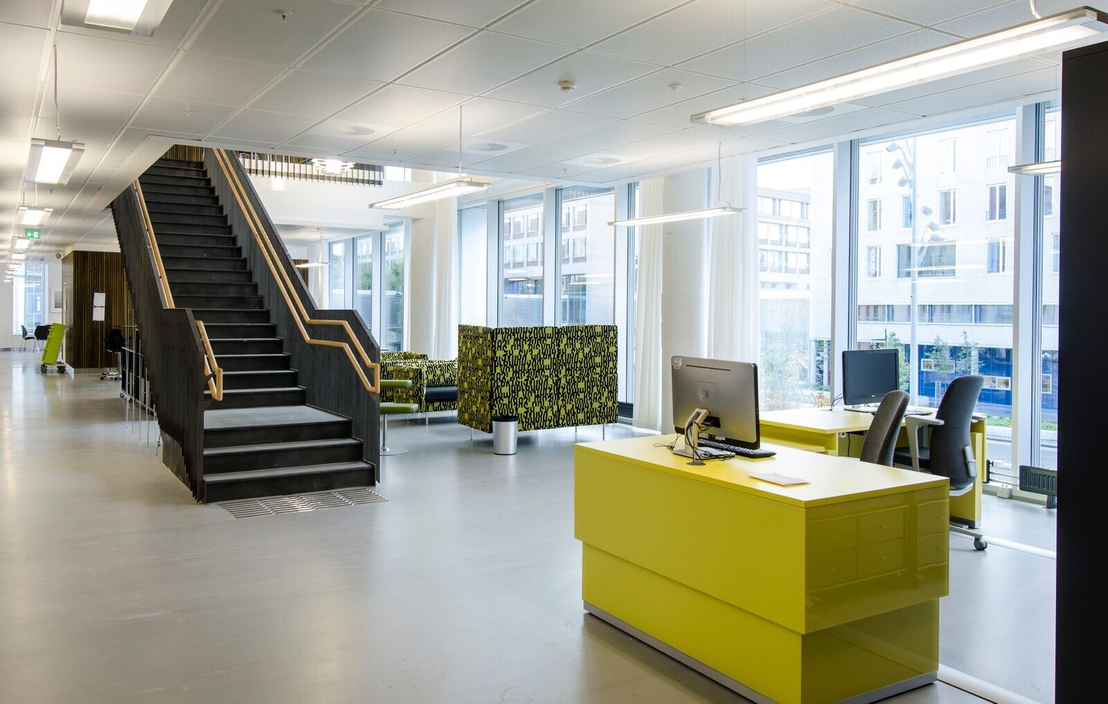 Interiør fra et sykehusbygg. En gul resepsjonspult står fremst i bildet. Bakover ses sittegrupper og store vinduer. En sort trapp leder opp til neste etasje.