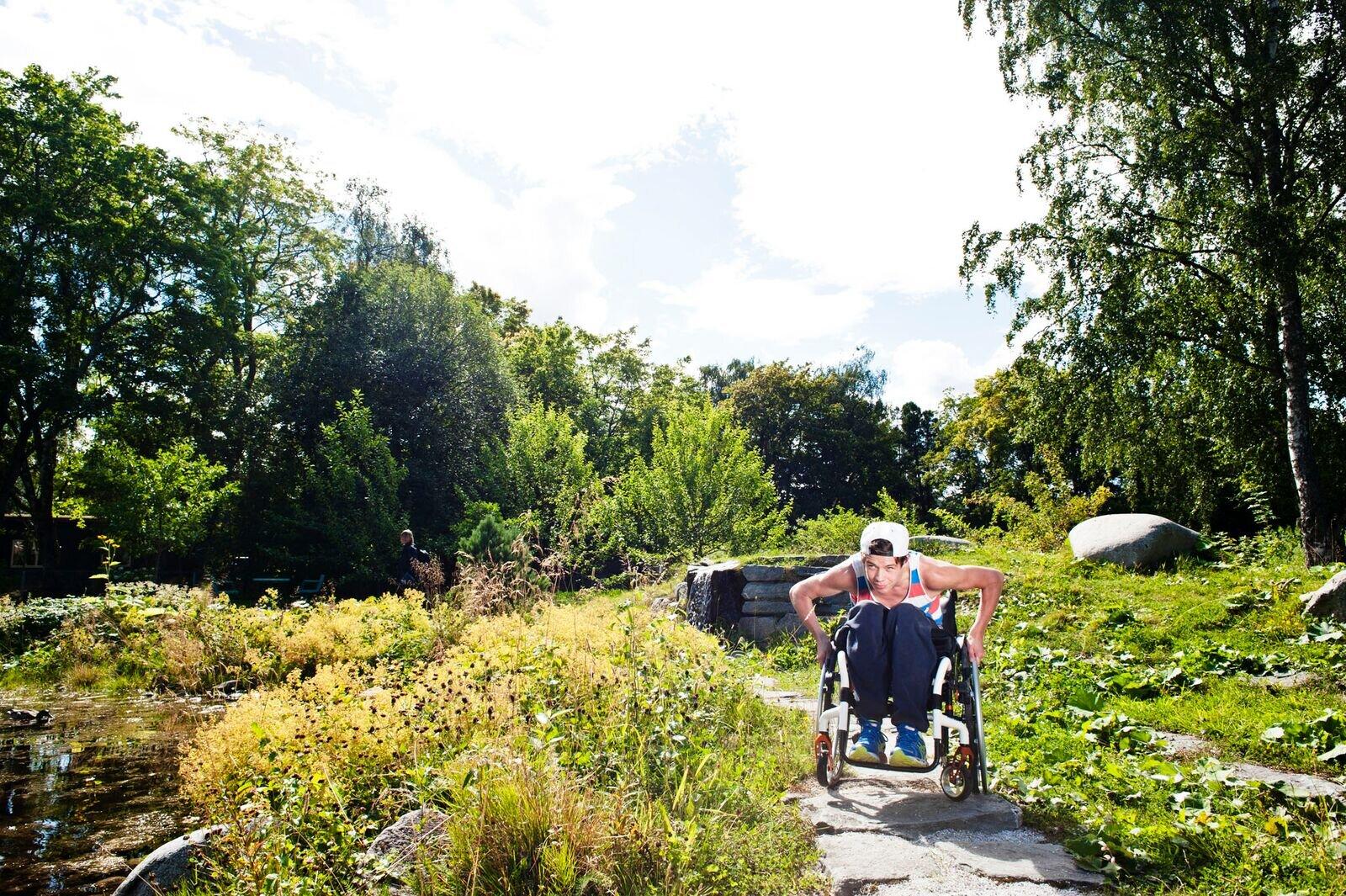 En ung mann i rullestol kjører på en ru steinsti gjennom et frodig beplantet område.
