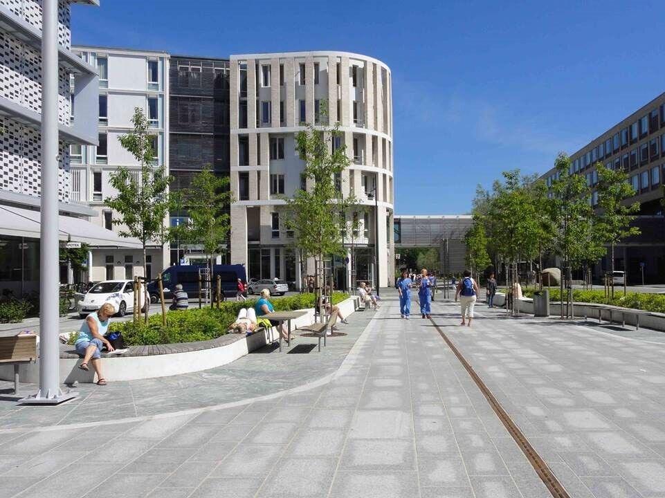 Moderne sykehusbebyggelse sett fra en bred gangvei gjennom området. Langs gangveien er det beplantning og sittegrupper, og flere mennesker som sitter og ligger i solen. Bygningsvolumene er varierte – ett buet i lys kledning og et lavere langt bygg på motsatt side.