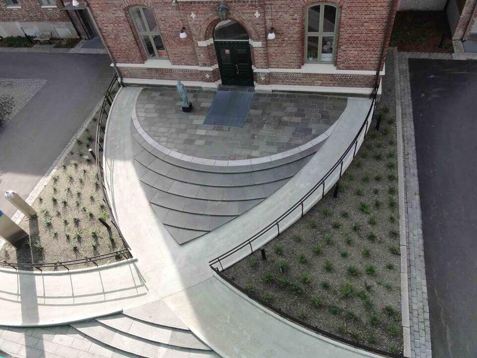 En symetrisk utformet adgangsplass foran et teglsteinskledd inngangsparti. Trinnfri adkomst i to akser som krysses, med trappetrinn i midten og beplantning på hver side.