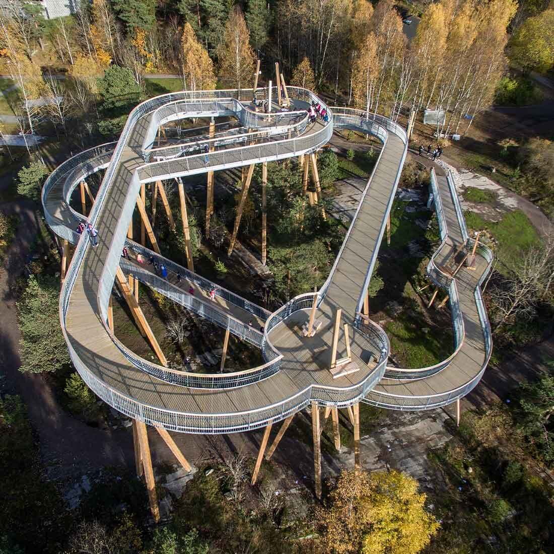 Fra fugleperspektiv: En rundløype hevet på massive trebjelker gir trinnfri ferdsel fra bakkenivå og til toppen av strukturen som en omkransert av løvskog.