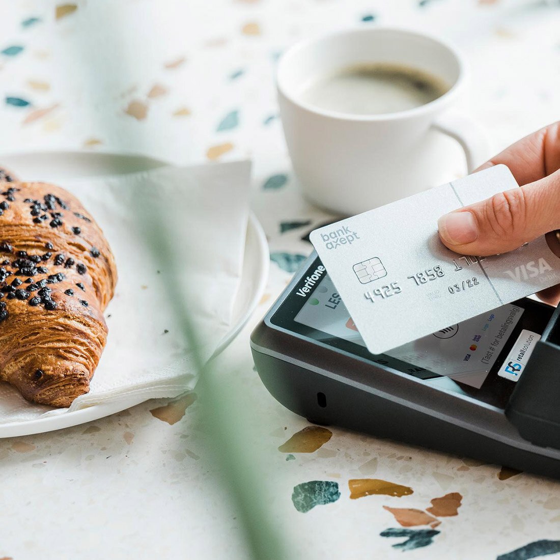 Nærbilde av et bankkort som "tæppes" mot en betalingsterminal – i en kafésetting.