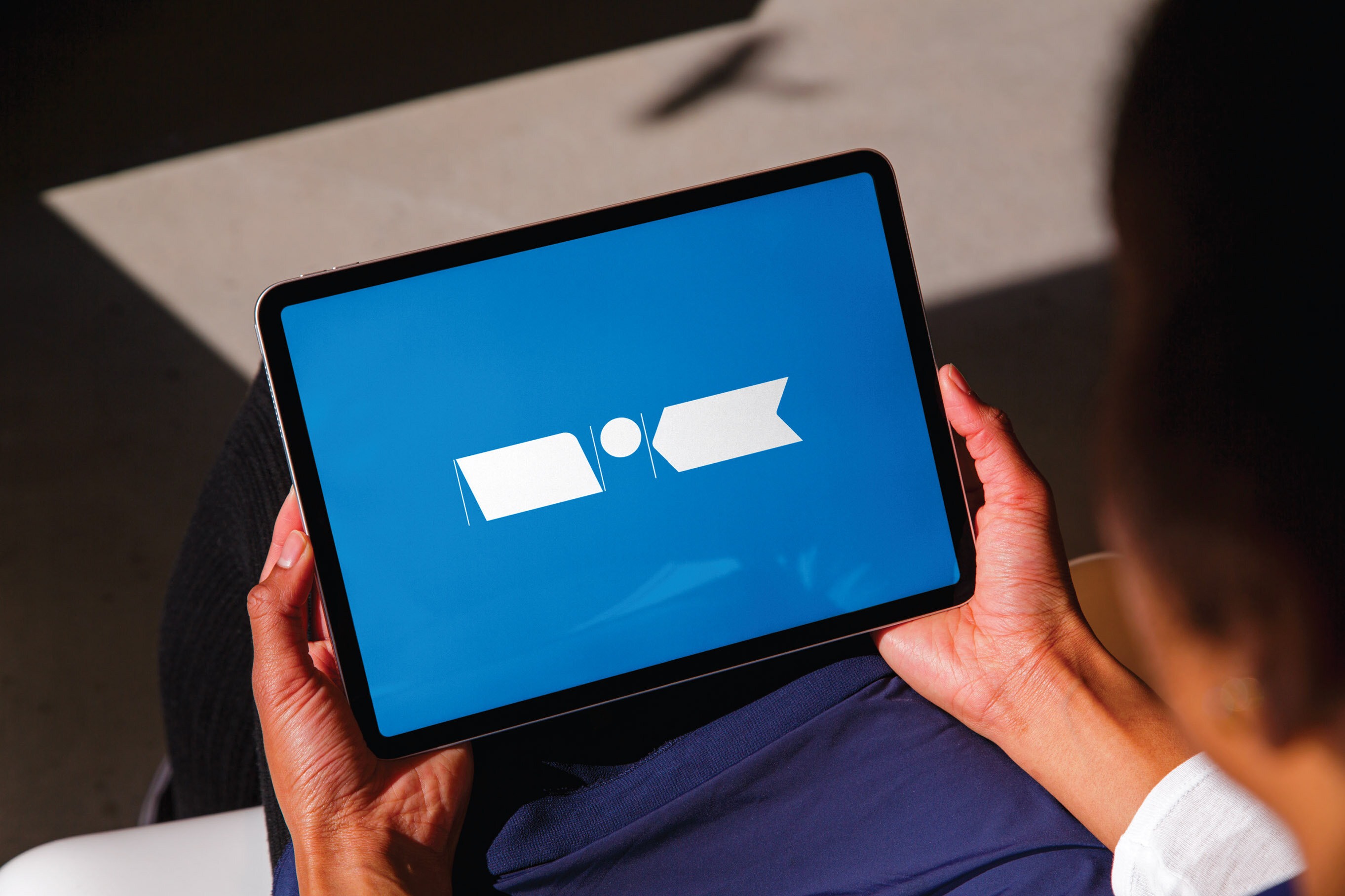 Hender som holder en iPad med hvit, strukket NRK-logo på mellomblå bakgrunn.