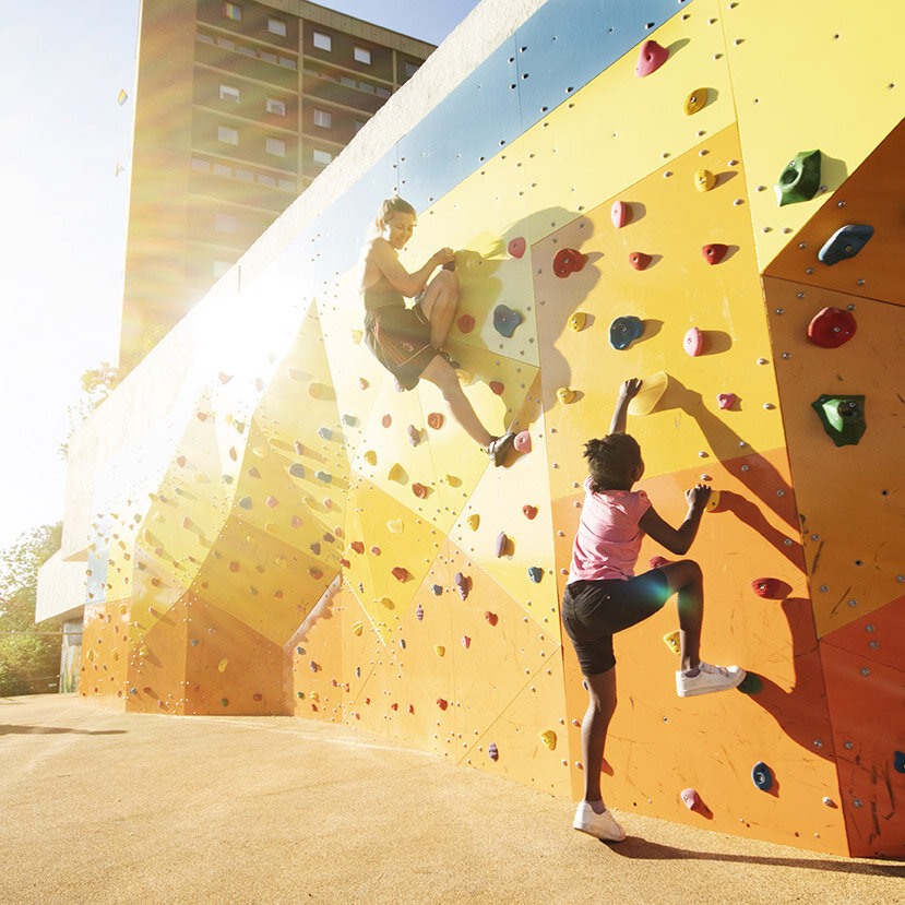 To jenter som klatrer i en gul klatrevegg i solskinnet.