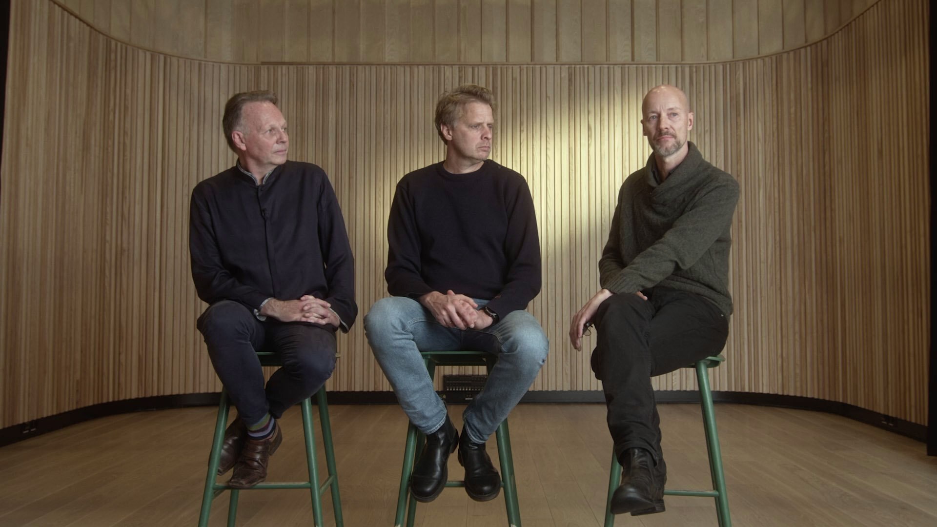 Tre menn sittende på krakker, som snakker sammen.