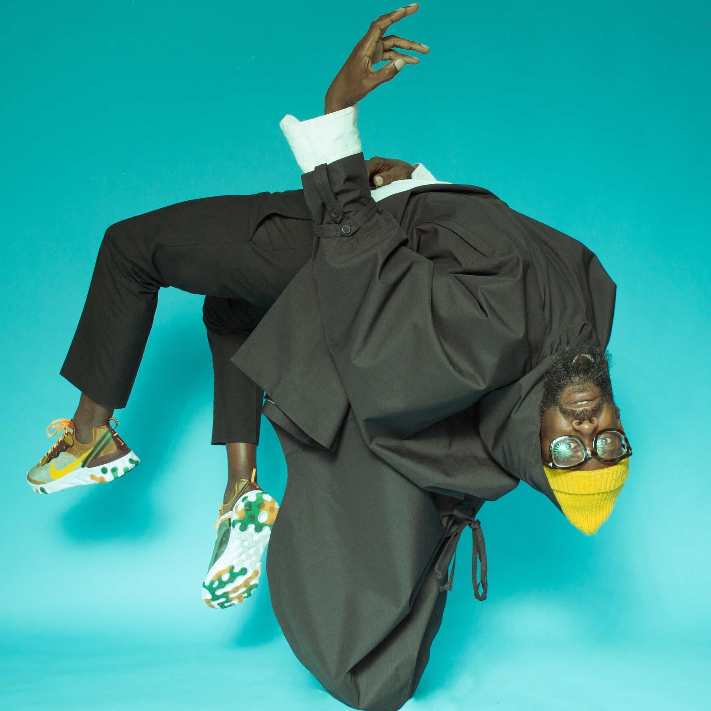 En mann i svart frakk og gul lue tar back-flip mot en turkisgrønn bakgrunn.