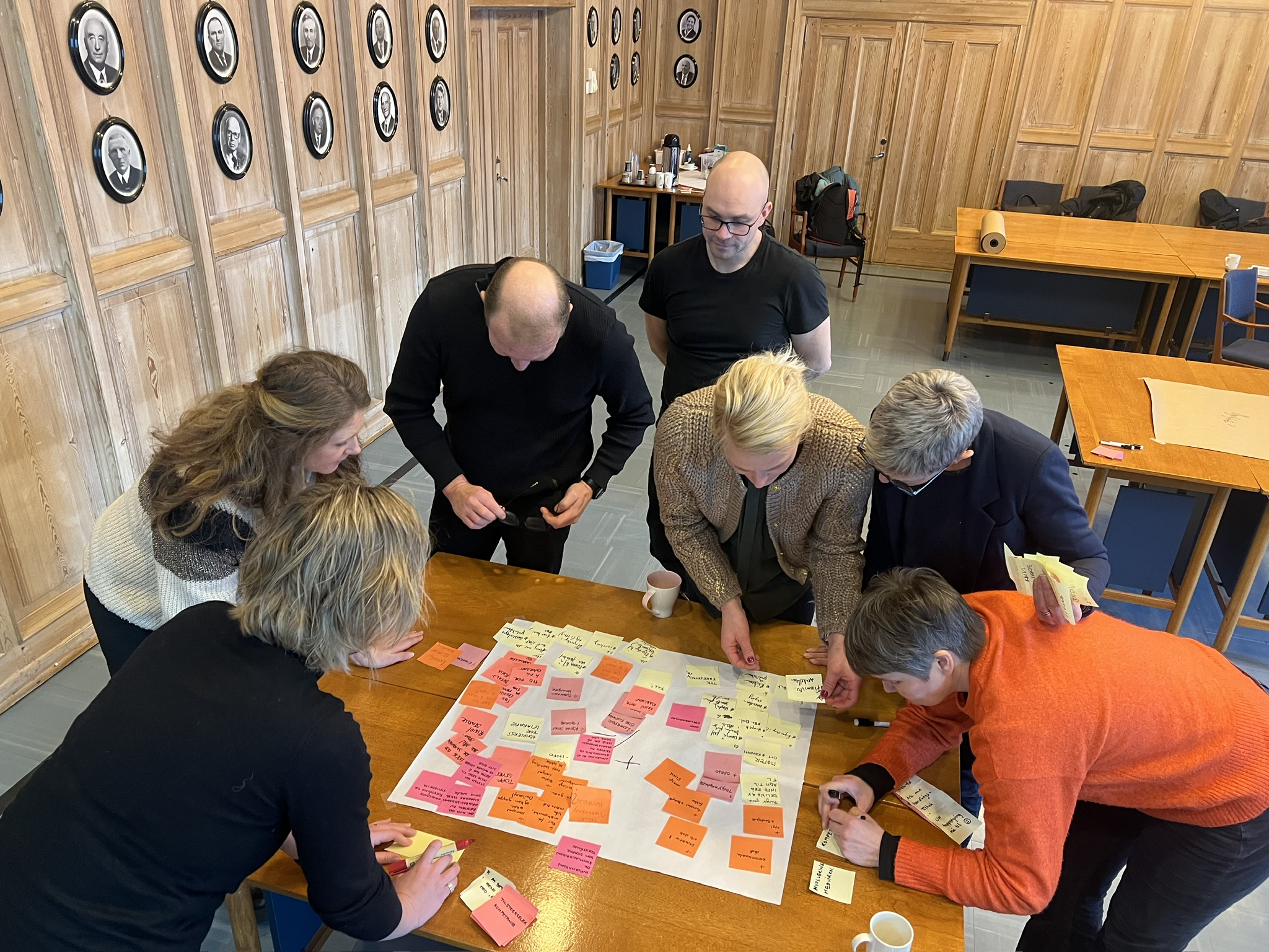 En gruppe mennesker står rundt et bord og ser på et stort ark fullt av fargerike post-it-lapper.