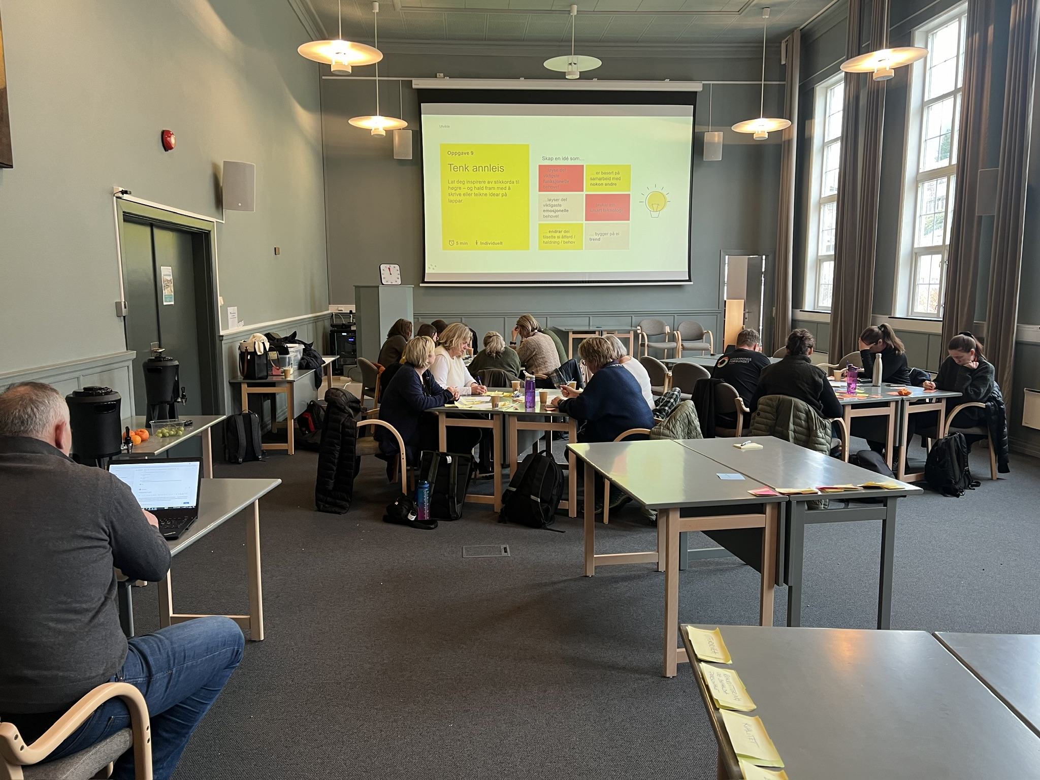 Mennesker sittende rundt bord i et stort lokale med en stor skjerm i enden.
