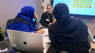 Foto av to damer med sjal som sitter med ryggen vendt mot kamera i samtale med en dame.