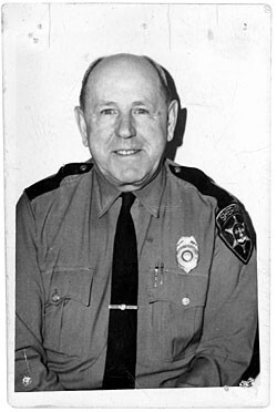 Dow Hover, an elderly bald white man wearing an officer uniform with badge.