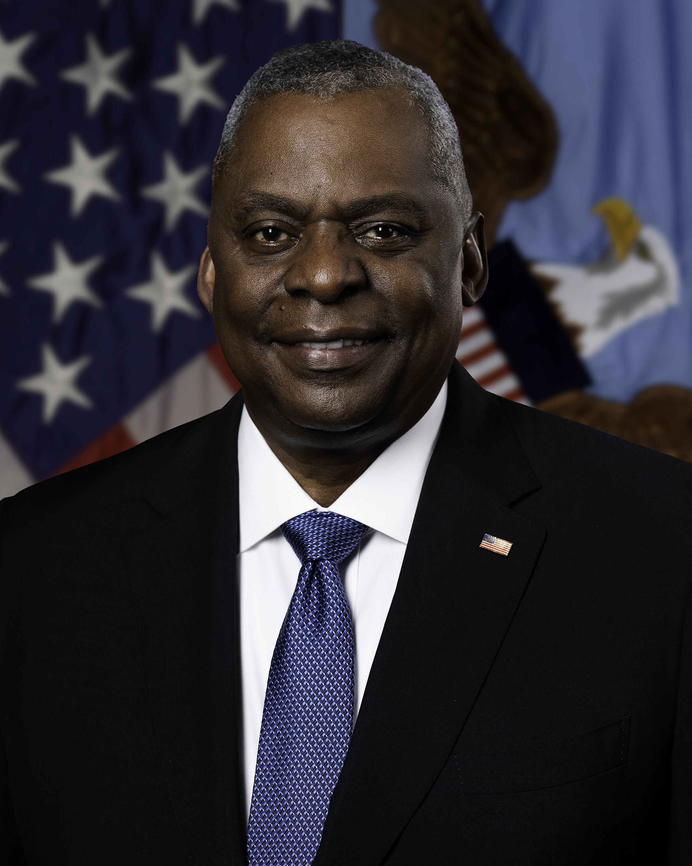Lloyd Austin, a Black man with gray hair in suit with blue tie and American flag pin in front of flag background