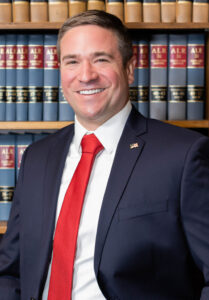 Andrew Bailey, a white man in a suit and red tie leaning against bookcase