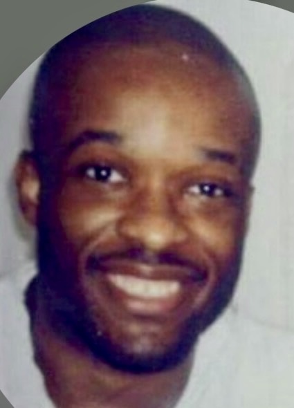 A smiling Black man with a shaved head and a light mustache and beard, wearing a white t-shirt.