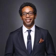 Wesley Bell, a Black man with glasses wearing a suit and tie in front of gray background