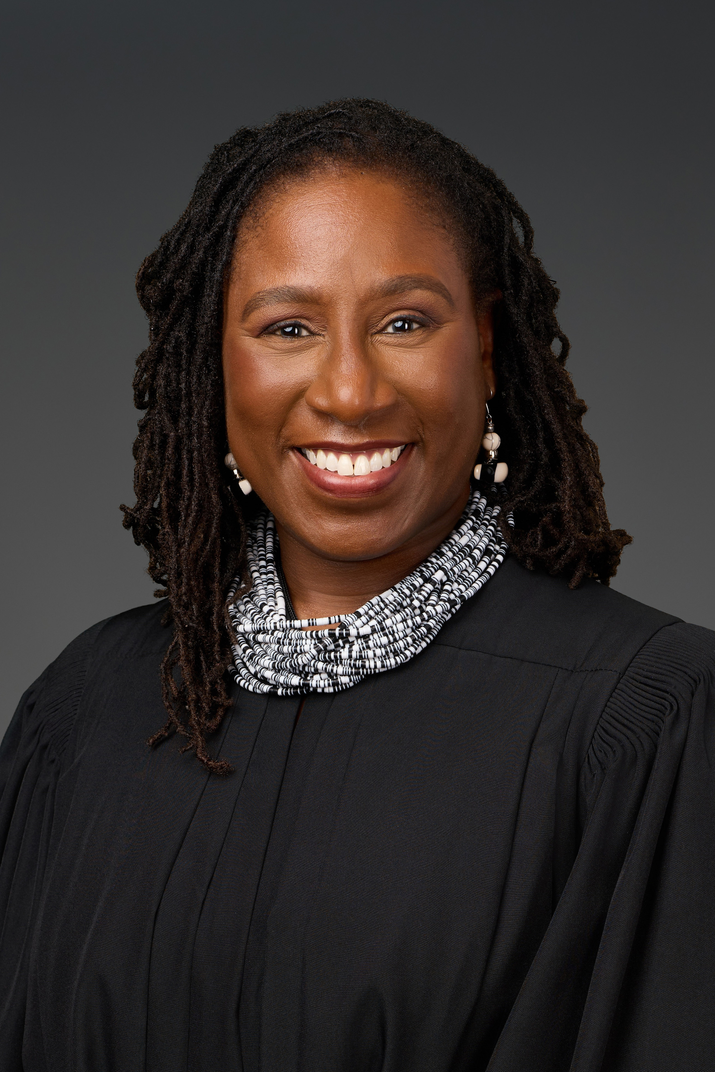 A smiling Black woman in judicial robes, a layered brown and white shell necklace, and ceramic earrings.